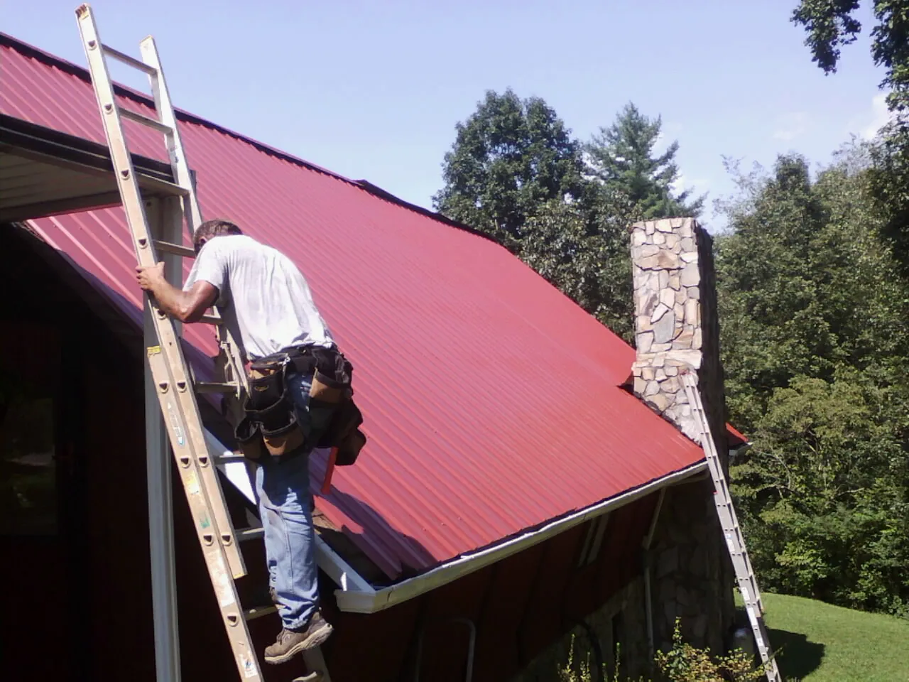 Roofer on a ladder beside a red metal roof and stone chimney — Soffit & Fascia Repair by Roofing Brothers in Kulpmont